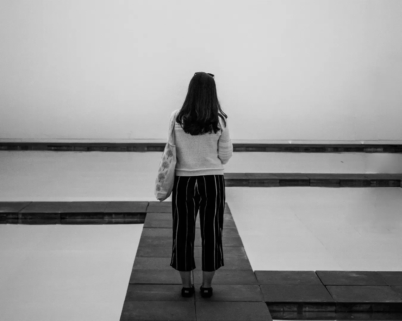 A woman stands alone in an open space, her posture calm and thoughtful. The scene captures a quiet moment of solitude, suggesting reflection, independence, or contemplation within a simple and uncluttered environment.