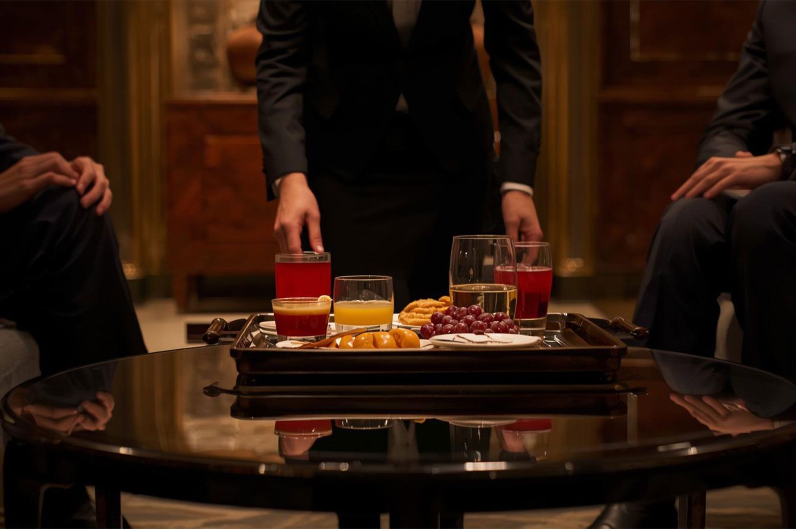 A formal waiter serving a tray of assorted juices, wine, and small appetizers (grapes, crackers, nuts) onto a glossy round table for seated business guests.