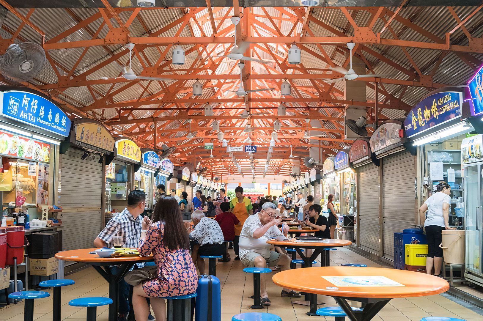 A wide-angle view inside a bustling Singaporean or Malaysian hawker center, showing the high, industrial roof structure, food stalls lining the walls, and patrons seated at orange tables with blue stools.