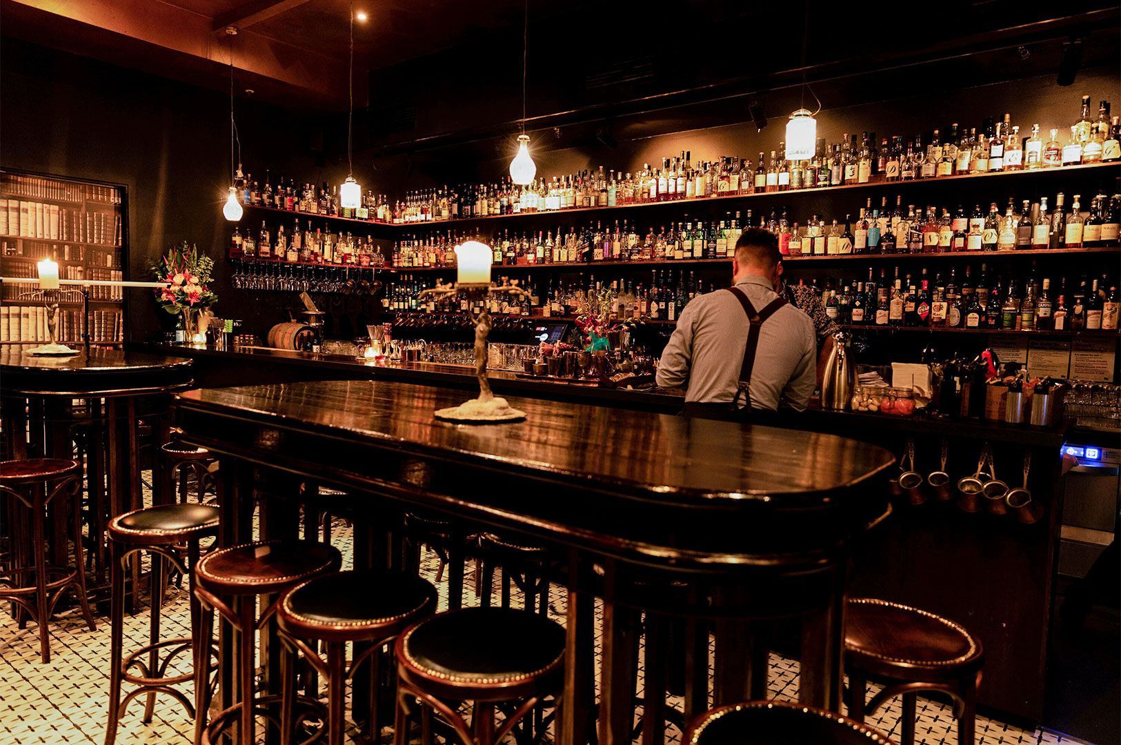 Atmospheric cocktail bar interior with a bartender working behind a long wooden counter, illuminated by hanging bulbs and a fully stocked liquor display.