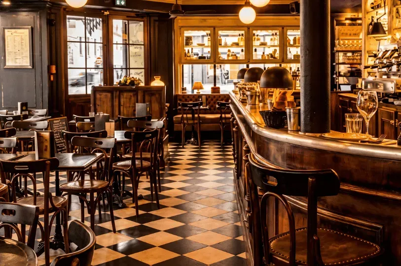 A cozy, softly lit bistro interior featuring a classic black-and-white checkered floor. The dining space is filled with dark wooden bistro chairs, small square tables, and a curved wooden bar counter, evoking a traditional and intimate European cafe atmosphere.