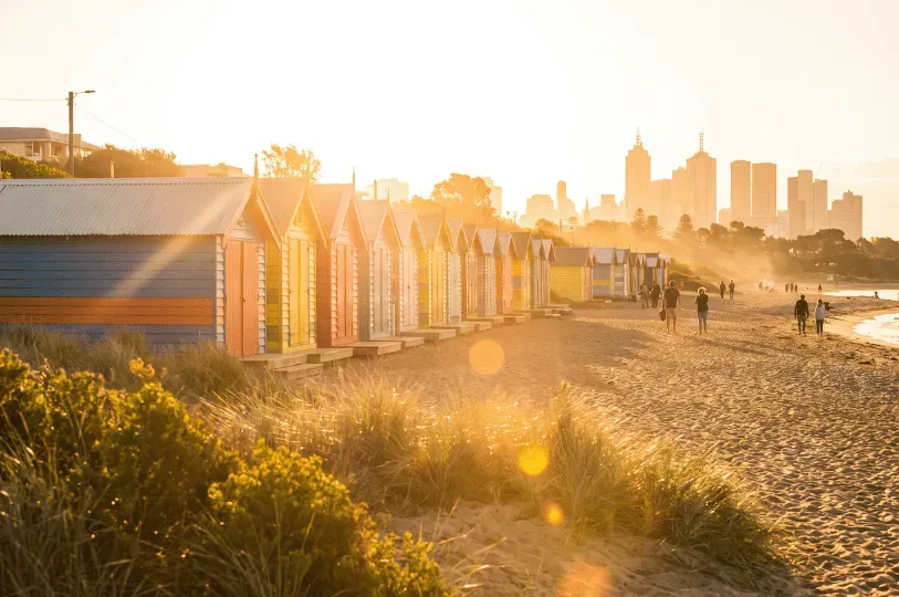 A row of colorful wooden bathing boxes along Brighton Beach at sunset, with golden light sweeping across the sand and the distant Melbourne city skyline visible in the background.