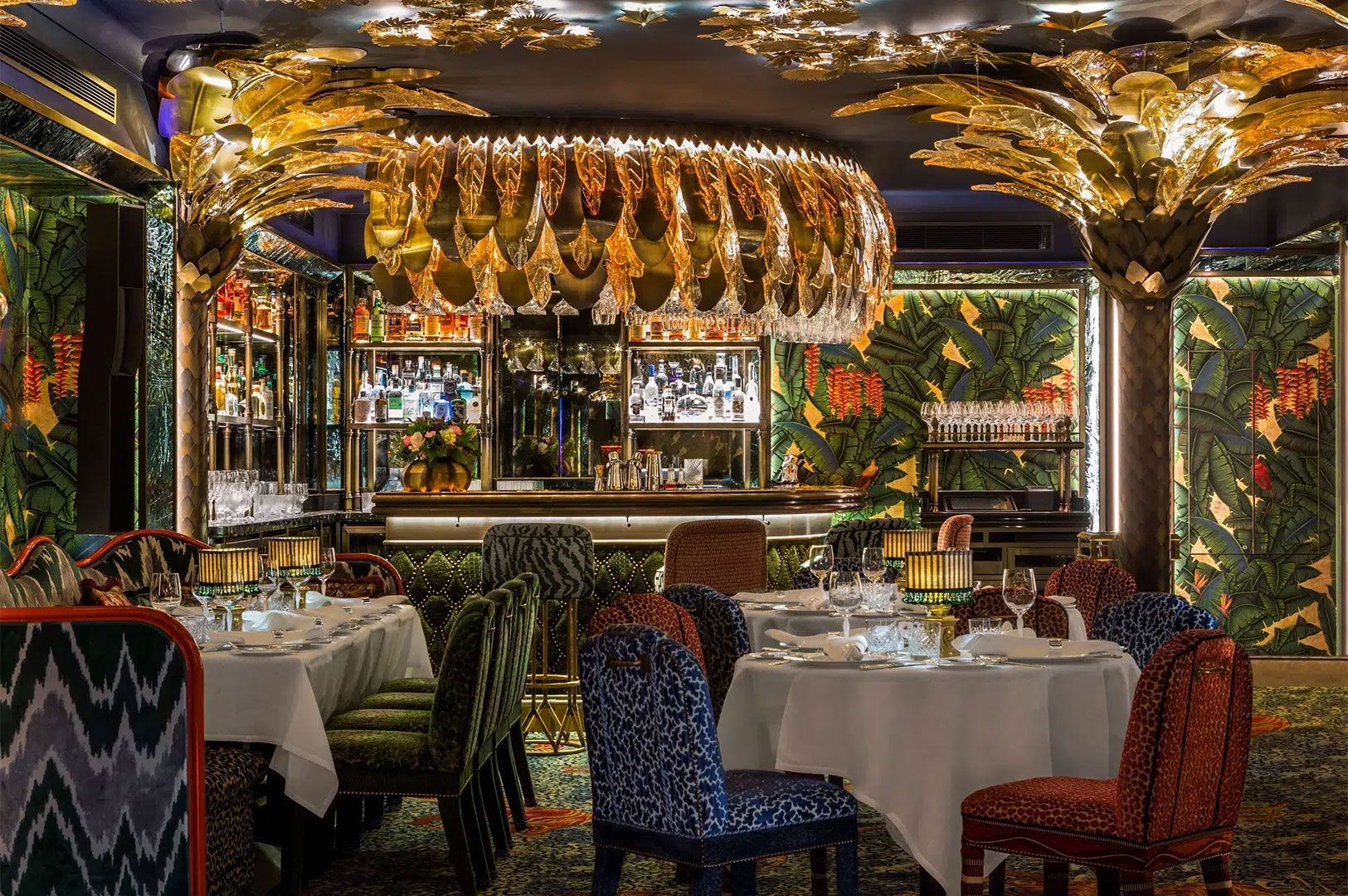 A lush, high-end restaurant and bar area with a tropical-themed decor, gold palm leaf accents on the ceiling, bold patterned seating, and a fully stocked bar.
