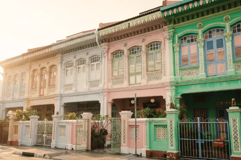 A row of ornate, pastel-colored heritage shophouses in shades of light blue, pink, and vibrant green. The traditional facades are decorated with intricate ceramic tiles, carved wooden window shutters, detailed plasterwork, and decorative metal front gates.