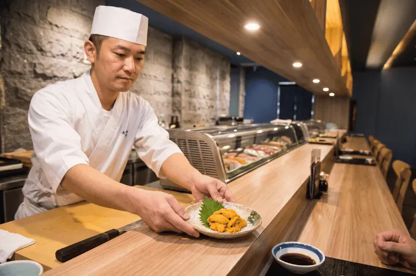 A sushi chef in a traditional white uniform carefully presenting a plate of fresh uni (sea urchin) across a pristine wooden omakase counter to a seated guest.