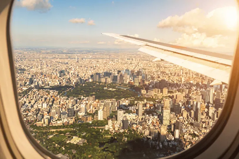 An aerial view of the dense Tokyo cityscape seen through an airplane window, highlighting a massive, lush green park surrounded by countless skyscrapers, with the airplane's wing visible against a bright, partly cloudy sky.