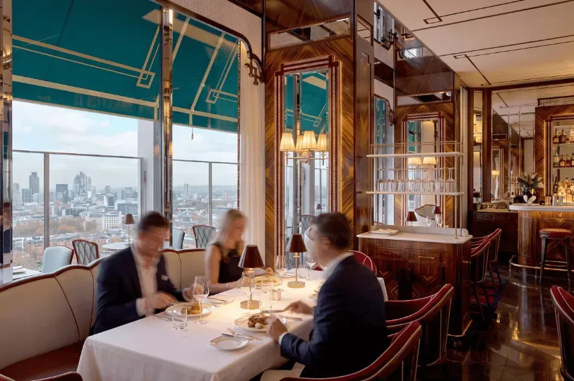 Diners captured in slight motion blur enjoying a meal at a white-tablecloth booth in a bright, Art Deco-inspired restaurant with polished wood paneling and large windows overlooking a city skyline.