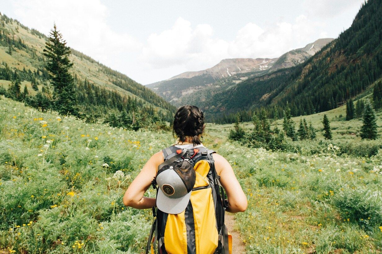 A woman wearing a bright yellow backpack hikes up a rocky mountain trail, surrounded by open sky and rugged peaks, capturing a moment of determination and outdoor adventure.