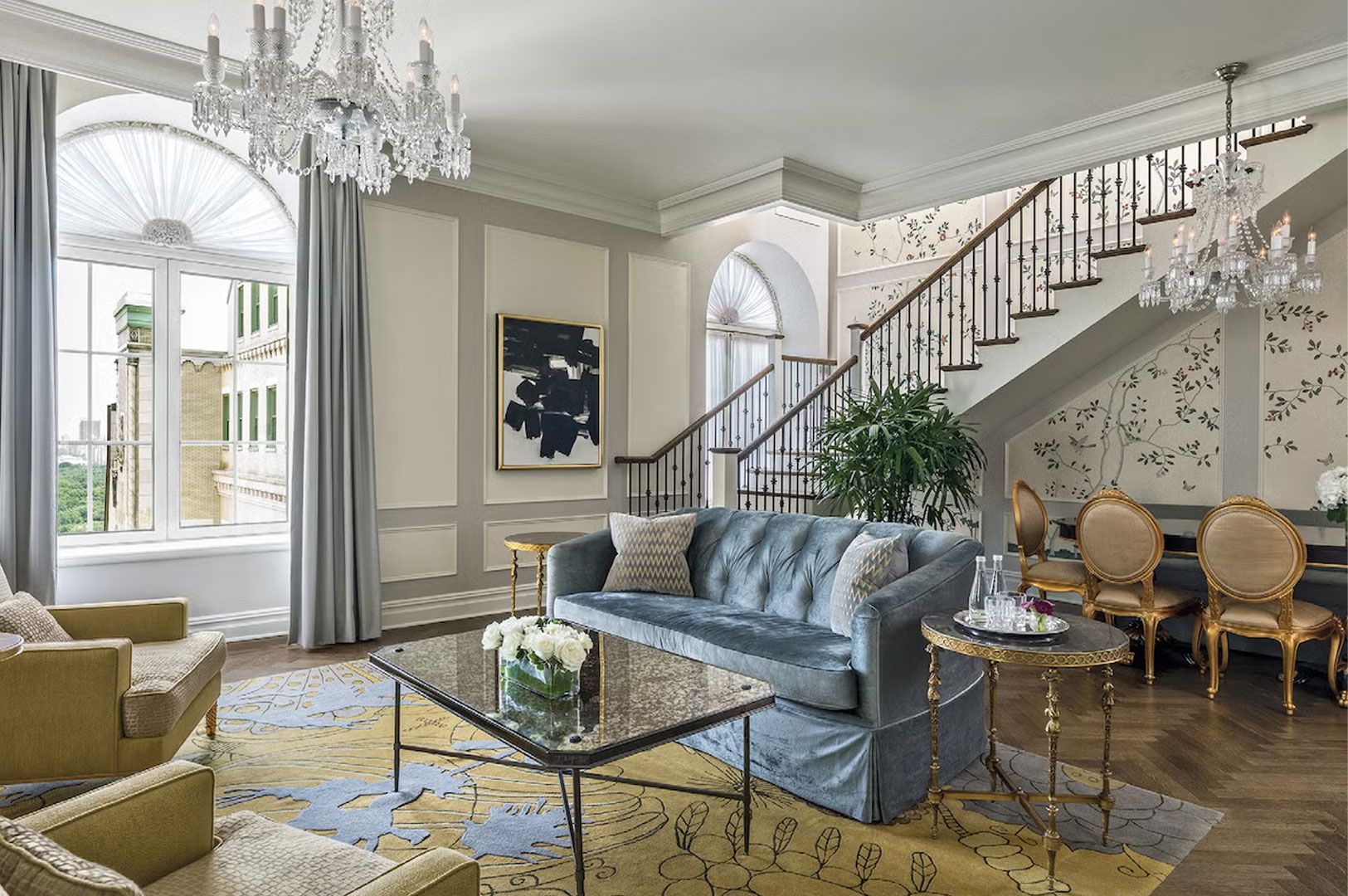 A grand, traditional hotel suite living room with a two-story layout, featuring a tufted blue sofa, crystal chandeliers, a winding staircase, and large arched windows.