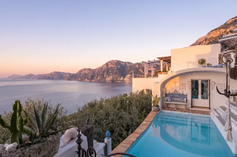 Soft morning light illuminates a white-washed villa terrace featuring a pristine turquoise plunge pool, arched Mediterranean architecture, and sweeping views of the rugged coastline.