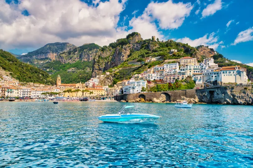 A bright blue speedboat drifts in crystal-clear waters before a picturesque Amalfi Coast town, its colorful, tightly packed buildings cascading down the steep, sun-drenched cliffs.