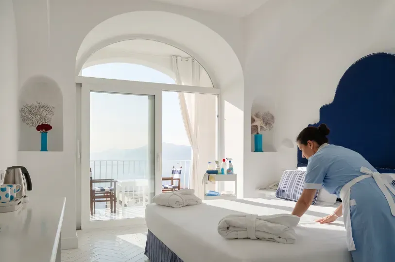 A housekeeper in a crisp blue uniform meticulously smooths the white linens of a bed inside a bright, arched room, with natural light streaming in from a balcony overlooking the sea.