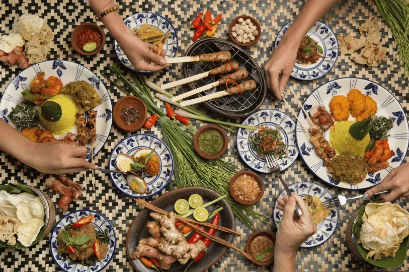 An overhead view of a group of people sharing a lavish, traditional Indonesian feast featuring yellow rice, satay, and fresh spices laid out on a woven mat.