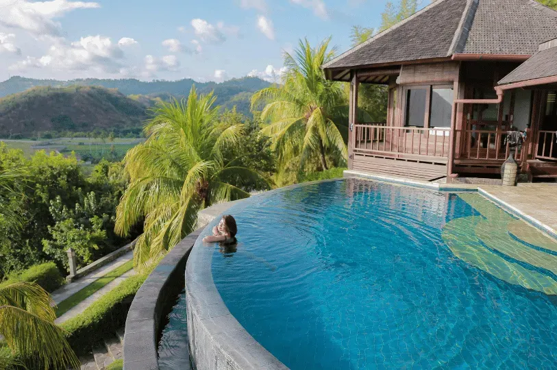 A woman relaxing at the edge of a curved infinity pool at a luxury villa, gazing out over a lush tropical valley and palm trees.