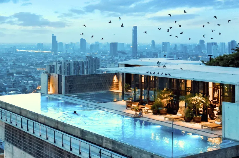 A luxurious, open-air penthouse with an infinity pool overlooking the dense, modern Bangkok skyline at dusk, with a flock of birds flying overhead.
