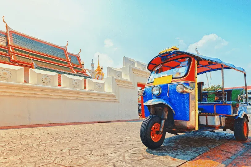 A vibrant blue and red traditional Thai tuk-tuk parked on a cobblestone street in front of the pristine white walls and ornate, sloping roofs of a Buddhist temple.