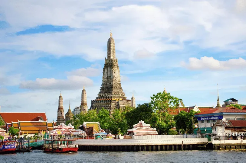 The iconic, intricately decorated spires of Wat Arun, the Temple of Dawn, rising majestically along the banks of the Chao Phraya River on a bright, cloudy day.