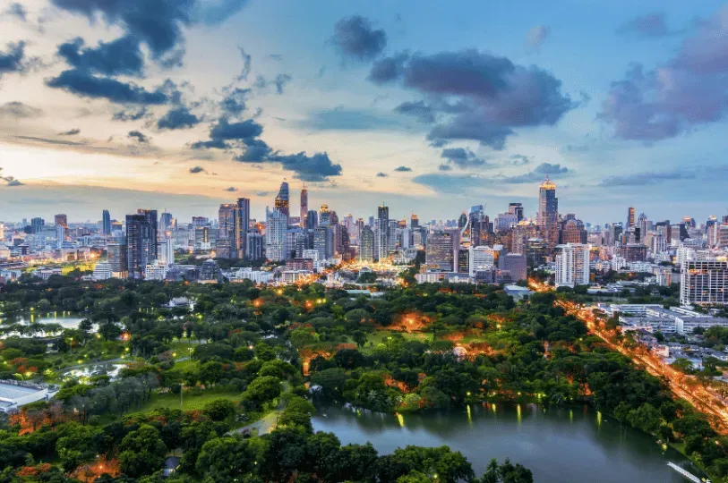 An expansive twilight view of Bangkok contrasting the lush, dark canopy and lakes of Lumphini Park in the foreground with a towering, illuminated skyline of modern skyscrapers in the background.