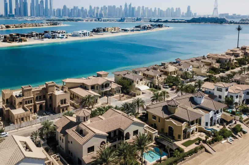 An aerial view of exclusive, sprawling luxury villas with private beaches along the vivid blue waters of a man-made island, juxtaposed against the hazy, dense Dubai skyline in the distance.