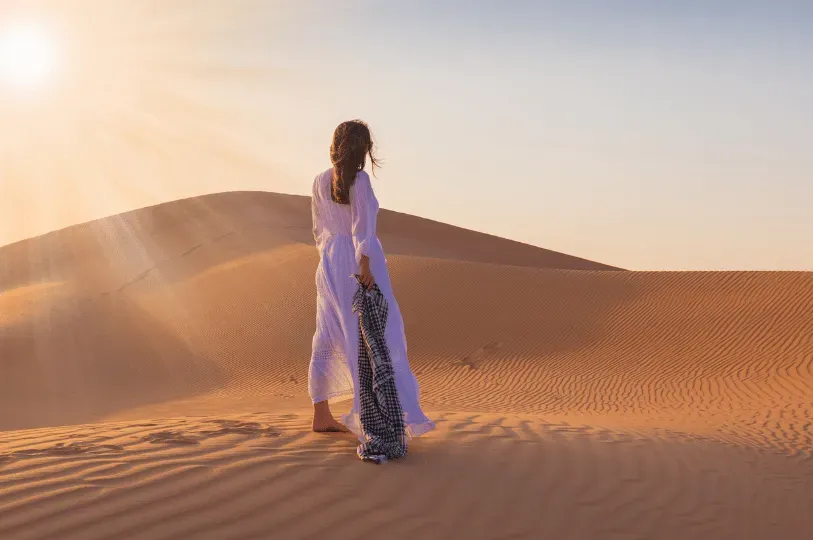 A woman in a flowing white dress stands alone looking out over vast, untouched orange sand dunes under a glaring sun, evoking the harsh, natural desert landscape that surrounds Dubai's manufactured oases.