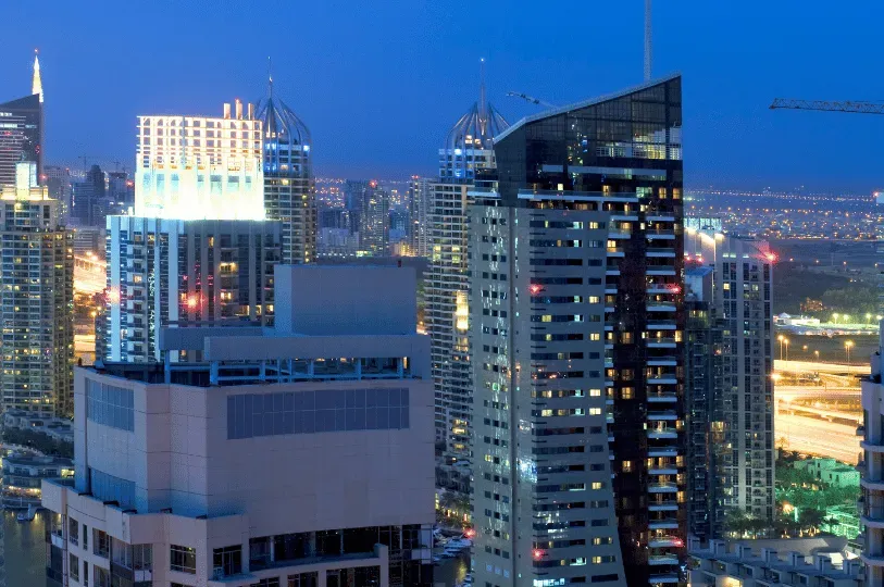A dense cluster of towering, illuminated skyscrapers in Dubai at twilight, highlighting the heavily developed, high-rise urban environment of the city.