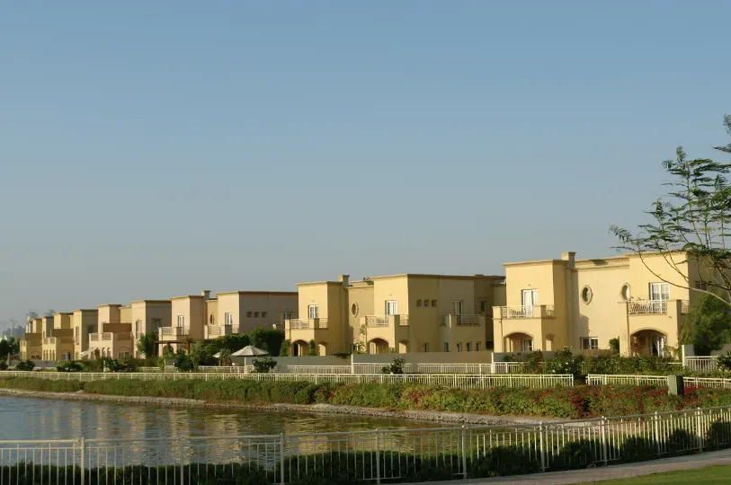 A uniform row of identical, closely-packed beige townhouses situated behind a fence along a waterfront promenade, illustrating the cookie-cutter density of some suburban residential developments in Dubai.