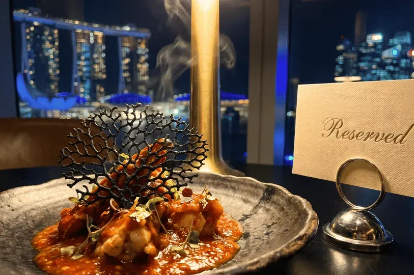 A close-up of an elegant fusion dish bathed in a rich sauce and topped with an intricate black coral-like tuile, placed next to a