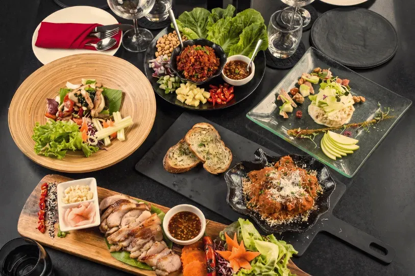 A top-down view of a diverse culinary spread featuring sliced roasted meats on a wooden board, vibrant Asian-inspired salads, artisanal bread, and rich curries arranged on a sleek dark dining table.