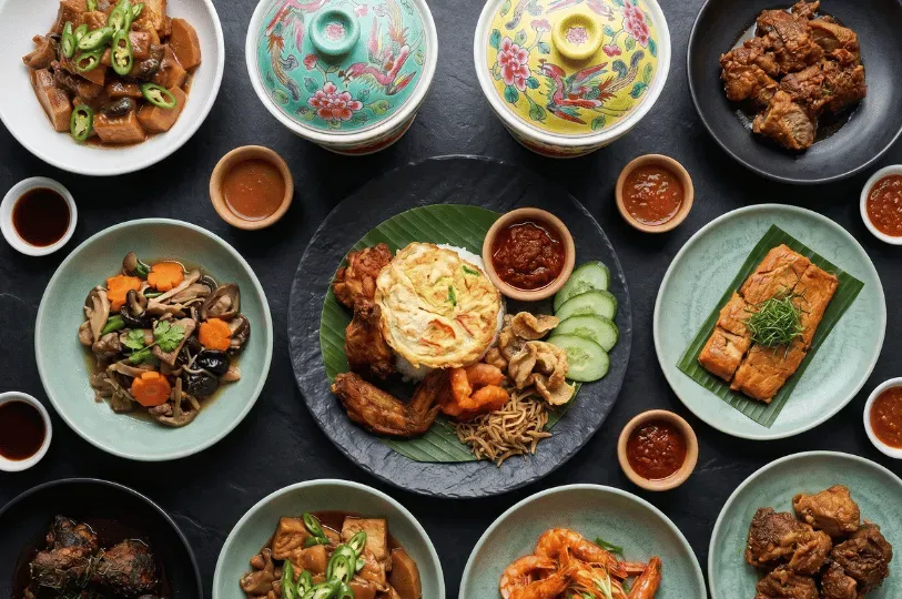 An overhead view of a vibrant, East-meets-West feast featuring a central rice dish with a fried egg, surrounded by beautifully plated braised meats, curries, and fresh prawns served in colorful ceramic bowls.