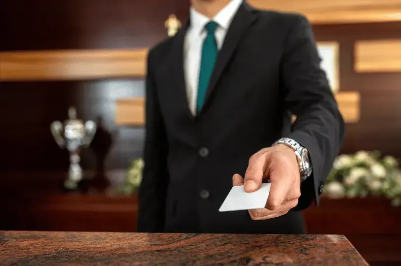 A close-up of a hotel staff member in a dark suit and green tie extending his hand to offer a white room key card across a granite reception desk.