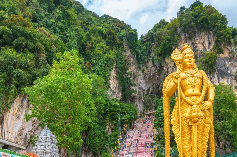 The iconic Batu Caves entrance, featuring a massive golden statue of Lord Murugan standing beside a steep, colorful staircase that leads up into a limestone cave covered in dense tropical greenery.