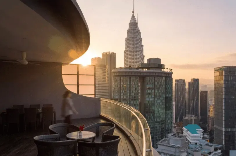 A blurred figure walks across a spacious, curved balcony of a luxury high-rise apartment furnished with dark wicker patio seating, overlooking a stunning sunset view of the Kuala Lumpur skyline and the Petronas Twin Towers.
