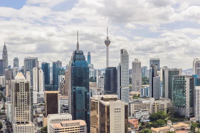 A daytime panoramic view of the diverse Kuala Lumpur cityscape, highlighting a dense collection of modern glass skyscrapers and the towering Menara KL (KL Tower) under a partly cloudy sky.