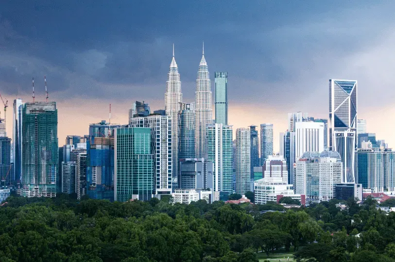A dramatic wide shot of the Kuala Lumpur skyline featuring the iconic Petronas Twin Towers surrounded by modern skyscrapers, with a vast, lush green park canopy in the foreground under a moody, overcast sky.