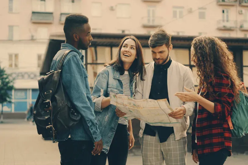 A diverse group of four young friends stand on a city street during the day, happily examining a map as they navigate Melbourne's streets in search of hidden venues.