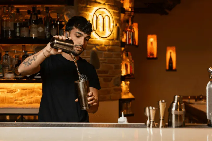 A bartender with a tattooed forearm skillfully pours a mixed cocktail from a silver shaker in a warmly lit bar featuring exposed brick and glowing wall alcoves.