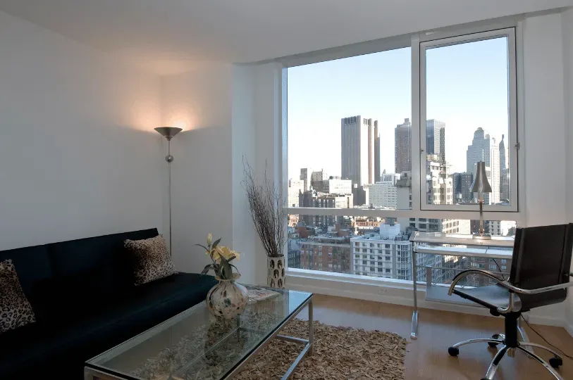A modern apartment interior featuring a living area and a sleek glass workspace positioned next to a large picture window, offering private, unobstructed views of neighboring city high-rises.