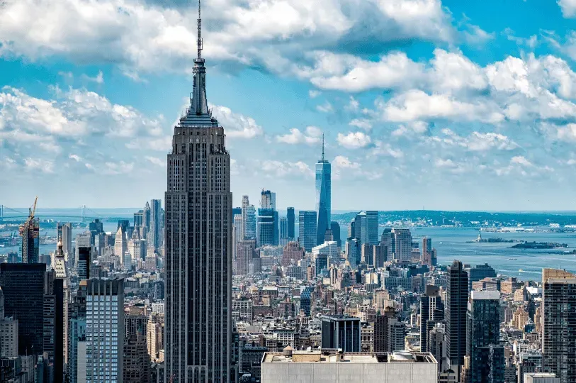 A dense daytime view of the New York City skyline under a bright, cloudy sky, prominently showcasing the architectural details of the Empire State Building with One World Trade Center in the distance.
