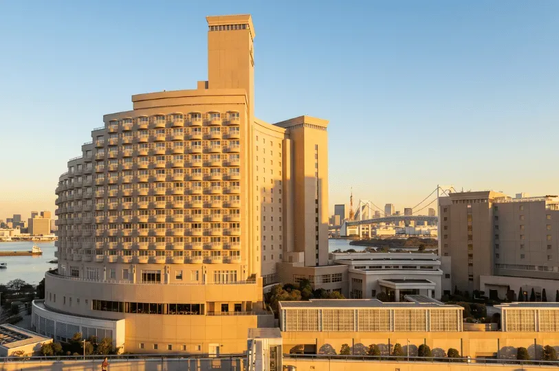 A grand luxury hotel with a curved, multi-story facade overlooking Tokyo Bay at sunset, with the Rainbow Bridge and Tokyo Tower visible in the serene background.