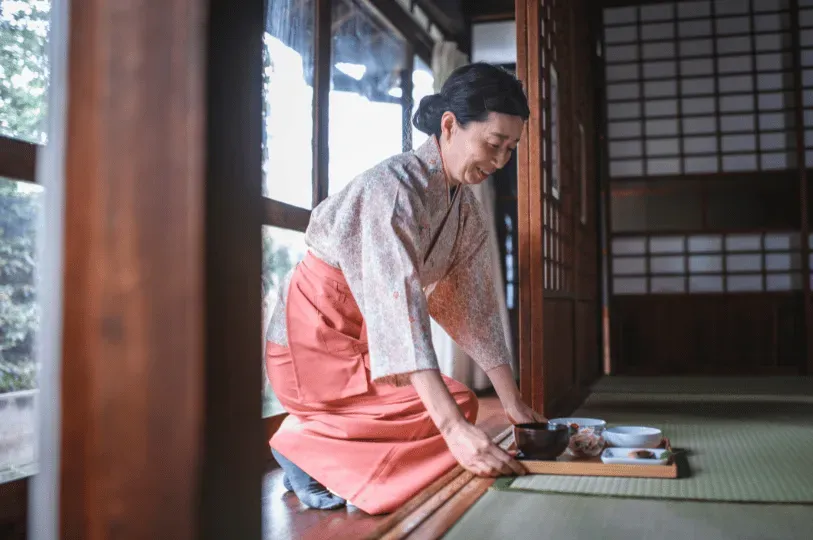 A smiling attendant in traditional Japanese attire kneeling gracefully on a tatami mat to serve a beautifully arranged in-room meal within a private, wood-paneled suite.