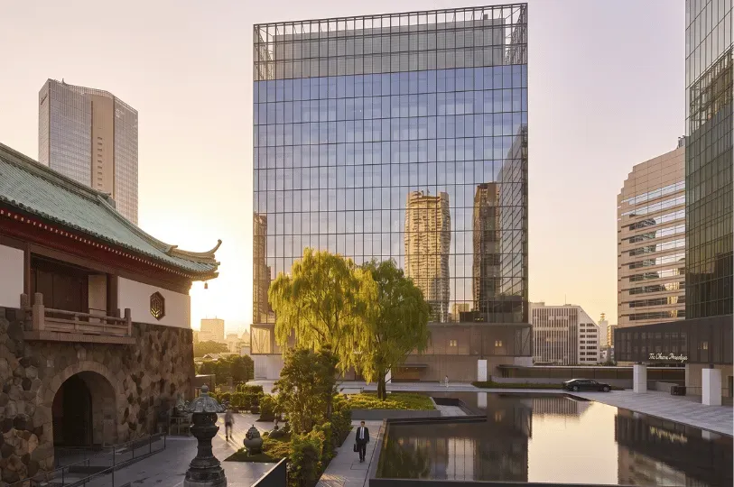 A tranquil courtyard featuring a still reflection pool that bridges a traditional Japanese stone gatehouse on the left with a towering, modern glass luxury hotel on the right.