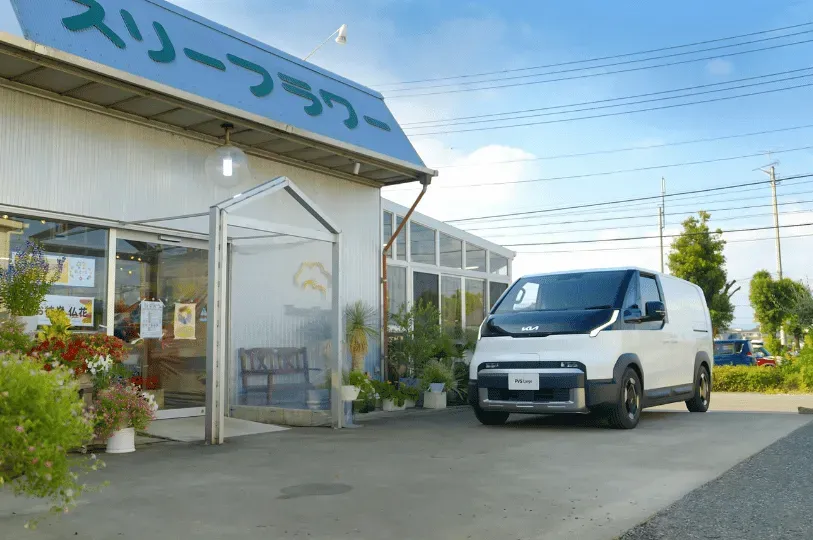 A modern white Kia electric cargo van parked discreetly outside a local Japanese flower shop with a blue awning and potted plants by the entrance.