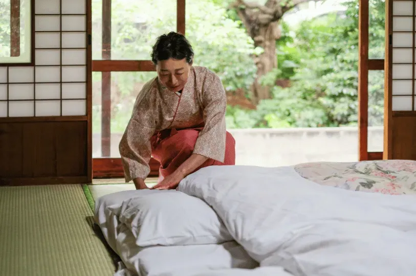 A dedicated attendant in traditional dress meticulously preparing a plush white futon bed on a tatami floor, with a peaceful, private green garden visible through open shoji doors.