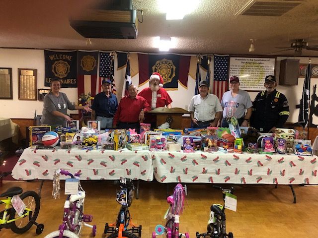 Seven people, including one dressed as Santa, stand behind tables filled with toys and bikes in a decorated meeting hall.