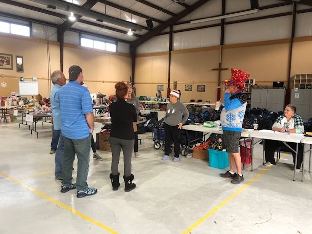 A group of people standing in a large, brightly lit community hall, gathered near tables and chairs for an indoor event.