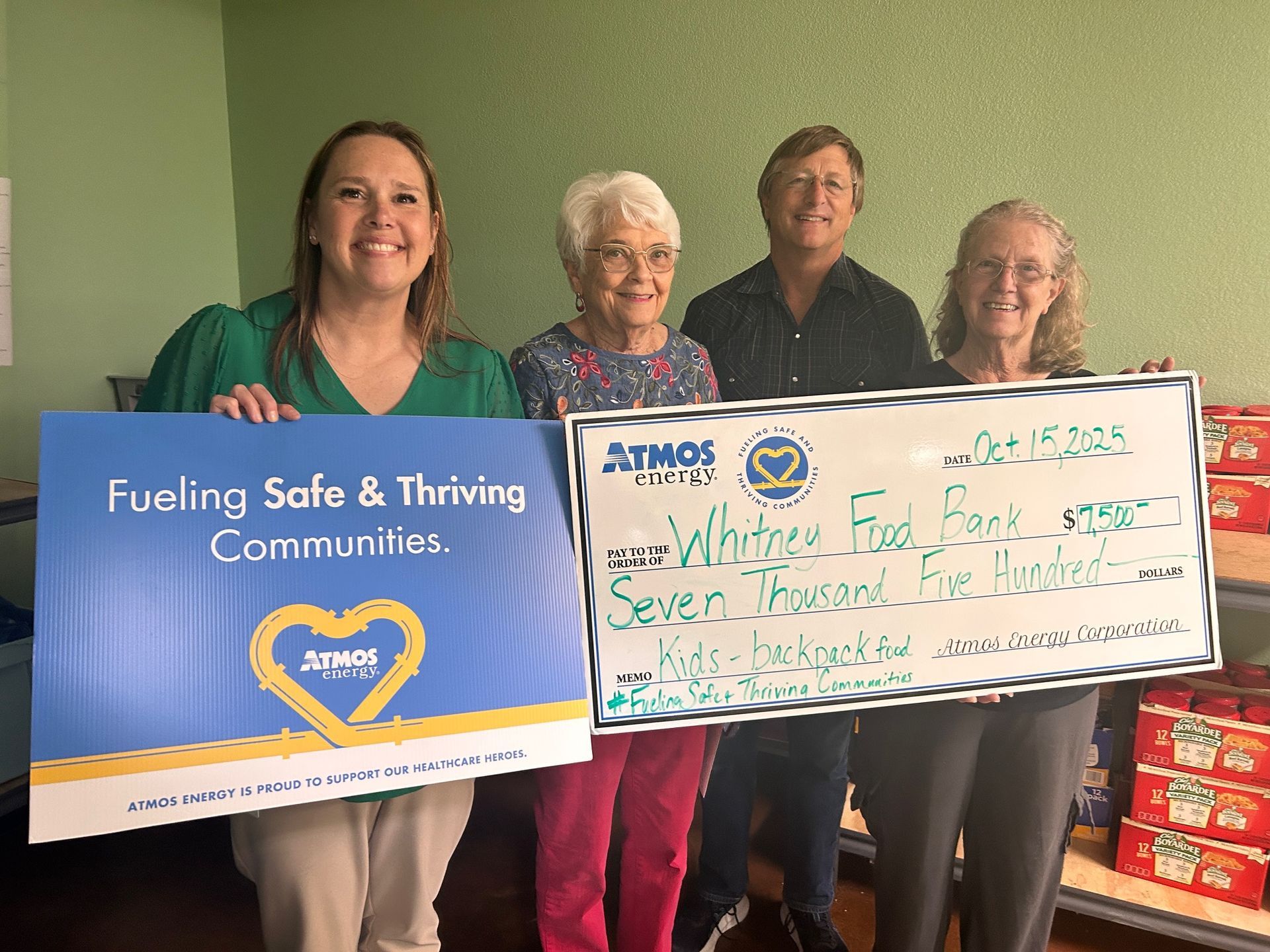 Four people hold a large Atmos Energy donation check for a food bank, smiling in a room filled with food supplies.