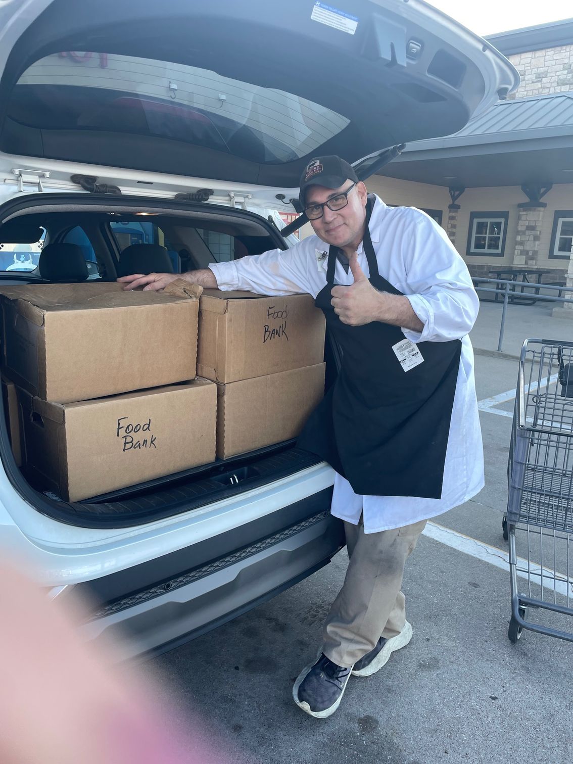 A person wearing a white shirt and black apron gives a thumbs-up while standing by a car trunk filled with cardboard boxes.