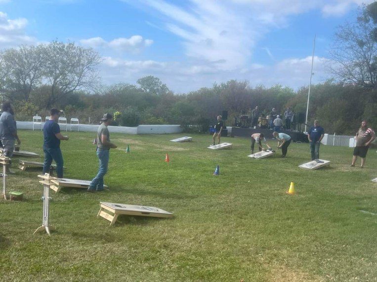 People playing cornhole on a grassy field under a sunny sky, with several game boards set up in rows.