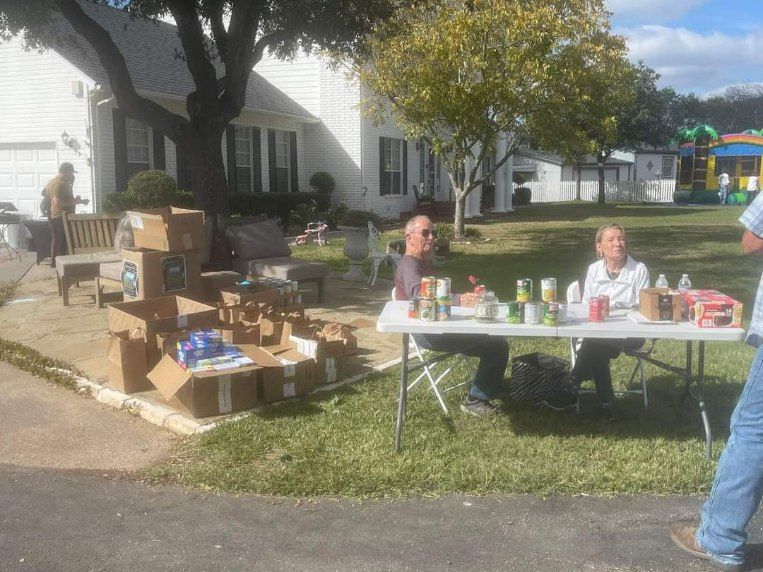People at a table with food items set up for donation on a sunny day. Boxes of items are on the ground.