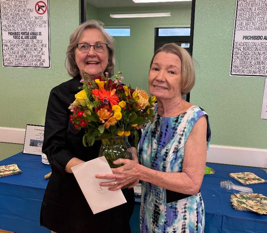 Two women smiling, one holding flowers and a card. Indoors, with a blue tablecloth and wall decor.